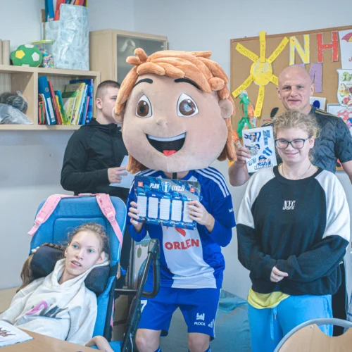 Na zdjęciu widać grupę osób w sali lekcyjnej, w tym osobę w kostiumie maskotki z dużą głową oraz dzieci, które pozują z uśmiechami i różnymi materiałami papierniczymi.