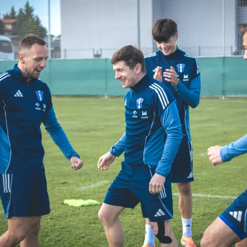 Grupa młodych piłkarzy w granatowych strojach przeprowadza trening na boisku, śmiejąc się i celebrując wspólnie spędzony czas.