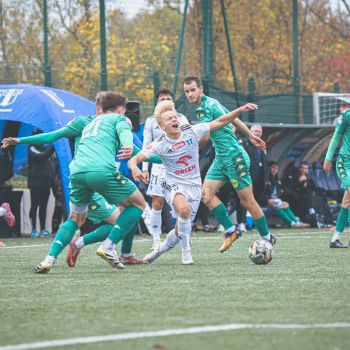 Na zdjęciu widać napiętą sytuację podczas meczu piłkarskiego, gdzie zawodnik w białym stroju stara się przejąć piłkę, otoczony przez rywali w zielonych strojach.