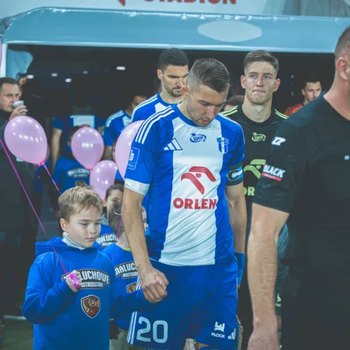 Na zdjęciu widoczny jest piłkarz w niebieskim stroju, trzymający się za rękę z dzieckiem, podczas gdy z tyłu widać zawodników i różowe balony, co sugeruje atmosferę radości i wsparcia przed meczem.