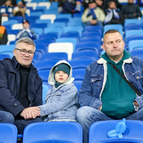 Na zdjęciu widoczni są mężczyzna i dziecko siedzący na trybunach, obaj ubrani w dżinsowe kurtki, a w tle widać innych kibiców.