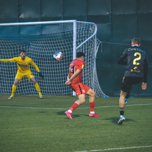 Na zdjęciu dwaj piłkarze rywalizują na boisku, podczas gdy bramkarz w żółtym stroju broni strzału na bramkę.