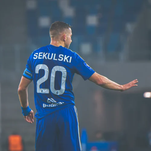 Na zdjęciu widoczny jest piłkarz w niebieskim stroju, z numerem 20 i nazwiskiem "SEKULSKI" na plecach, który gestykuluje na boisku w trakcie meczu.