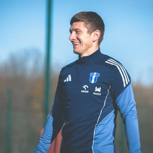 Na zdjęciu widoczny jest uśmiechnięty mężczyzna w sportowej odzieży, stojący na boisku w słoneczny dzień.