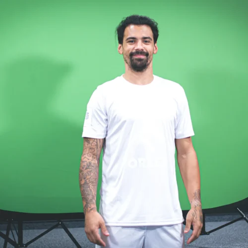 Mężczyzna o tatuażach stoi na tle zielonego ekranu, uśmiechając się i nosząc biały t-shirt.