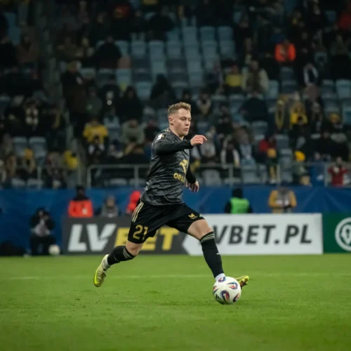 Na zdjęciu widoczna jest akcja piłkarska, w której zawodnik w czarnym stroju dąży do piłki, a jego przeciwnik w żółtej koszulce jest niewiele dalej.