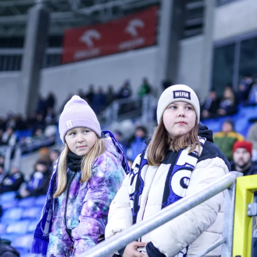 Dwie dziewczynki w ciepłych ubraniach kibicują na stadionie, otoczone przez tłum widzów.