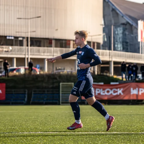 Młody piłkarz w niebieskim stroju z radością wskazuje na coś podczas meczu na boisku, w tle widać kibiców oraz budynki.