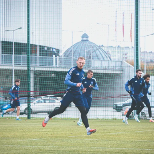 Na zdjęciu widocznych jest kilku piłkarzy podczas treningu na boisku, biegających w jednolitych strojach sportowych.