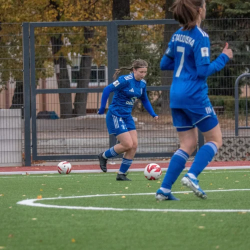 Na zdjęciu widać dwie piłkarki w niebieskich strojach, grające na boisku piłkarskim, podczas treningu w jesiennej scenerii.