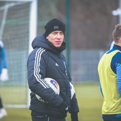 Na zdjęciu widać trenera piłkarskiego z piłkami, rozmawiającego z zawodnikiem podczas treningu na boisku.