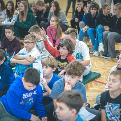 Na zdjęciu widać grupę dzieci i nastolatków siedzących na podłodze w sali, z jedną osobą podnoszącą rękę, co sugeruje aktywny udział w zajęciach.