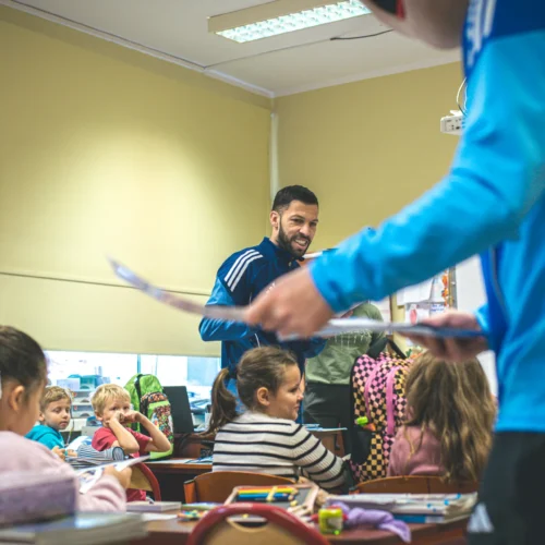 Zdjęcie przedstawia nauczyciela w niebieskiej bluzie, który uśmiecha się do uczniów siedzących przy biurkach w klasie.