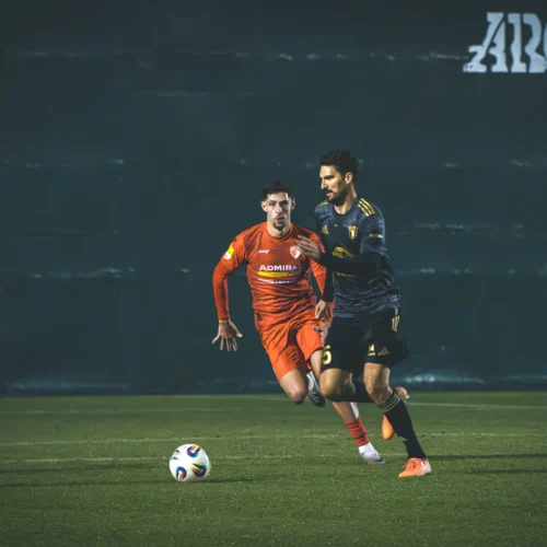 Na zdjęciu dwóch piłkarzy biega po boisku, a jeden z nich w czerwonym stroju goni drugiego w czarnym, który kontroluje piłkę.