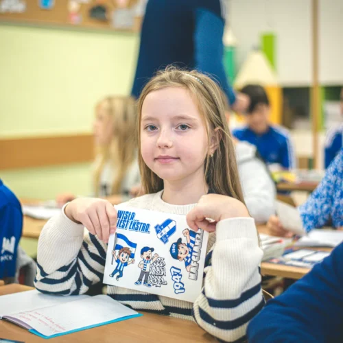 Dziewczynka w klasycznym swetrze trzyma kartkę z grafiką, uśmiechając się, podczas lekcji w szkole, a w tle widać innych uczniów.