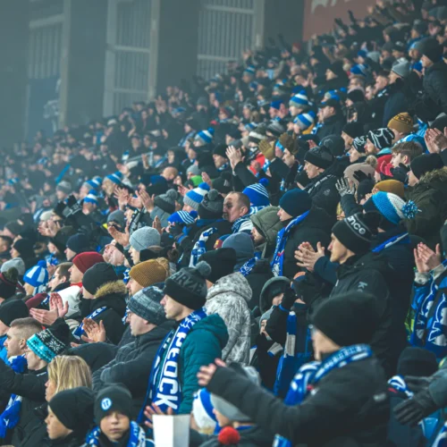 Tłum widowni na stadionie, w którym kibice w zimowej odzieży i szalikach głośno dopingują swoją drużynę.