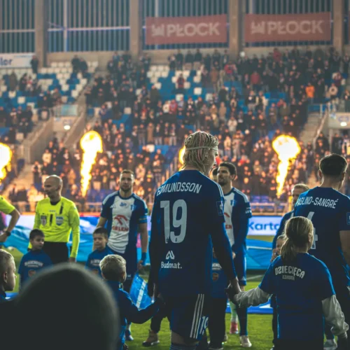 Na zdjęciu widoczni są piłkarze oraz dzieci trzymające się za ręce, podczas ceremonii przedmeczowej na stadionie, z tłumem widzów w tle i efektami pirotechnicznymi.