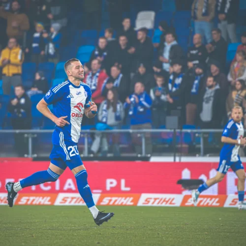 Na zdjęciu widać piłkarza w niebiesko-białym stroju biegnącego po boisku, a w tle znajdują się wiwatujący kibice.