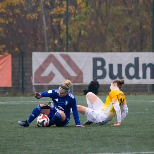 Na zdjęciu widoczna jest sytuacja z meczu piłkarskiego, w której dwie zawodniczki, jedna w niebieskim stroju, a druga w żółto-białym, leżą na boisku po starciu.