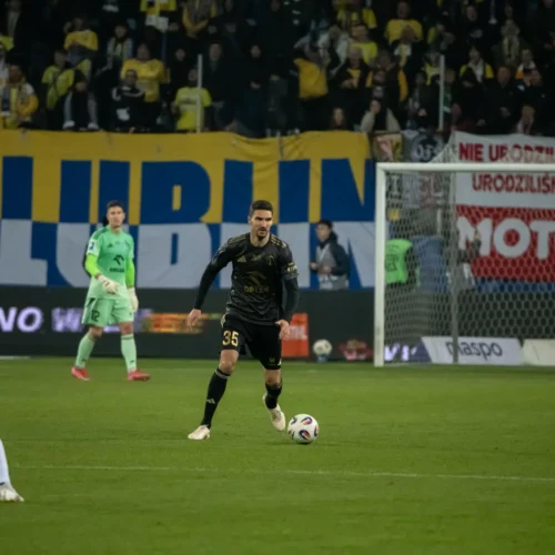 Na zdjęciu widoczna jest akcja na boisku piłkarskim, w której zawodnik w czarnej koszulce dynamicznie zmierza w kierunku piłki, a w tle na trybunach widać kibiców z dużą flagą i transparentami.