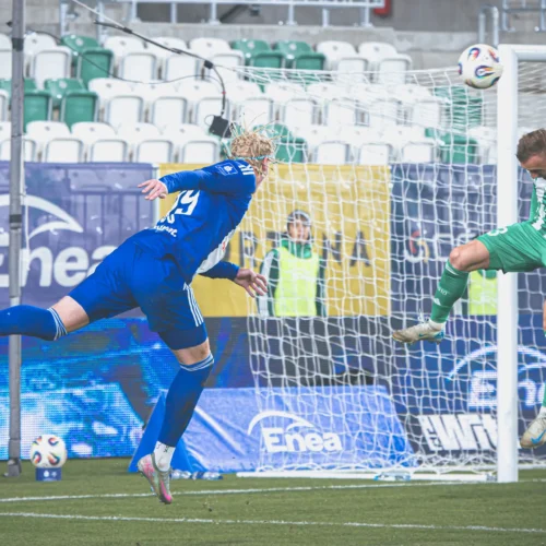 Na zdjęciu widoczna jest dynamiczna sytuacja z meczu piłkarskiego, gdzie zawodnik w niebieskim stroju strzela do bramki, a bramkarz w zielonym stroju stara się obronić strzał.