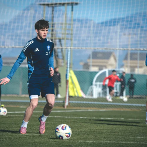 Na zdjęciu widać młodego piłkarza trenującego na boisku, podczas gdy w tle znajdują się inni zawodnicy i elementy infrastruktury sportowej.