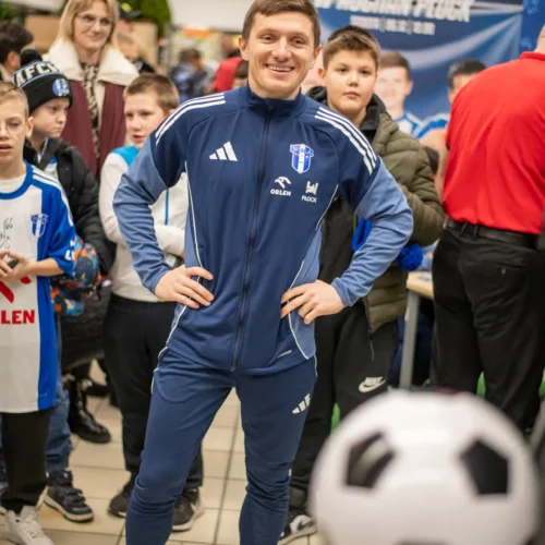 Na zdjęciu widać uśmiechniętego mężczyznę w niebieskim stroju sportowym, który stoi wśród grupy dzieci podczas wydarzenia promującego piłkę nożną.