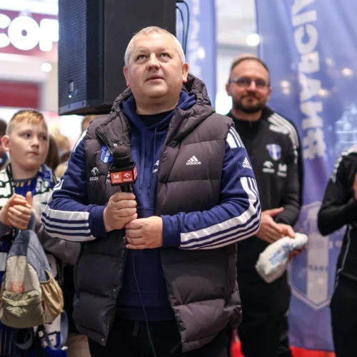 Mężczyzna trzyma mikrofon i patrzy w górę, otoczony przez grupę ludzi w tle, podczas wydarzenia w centrum handlowym.