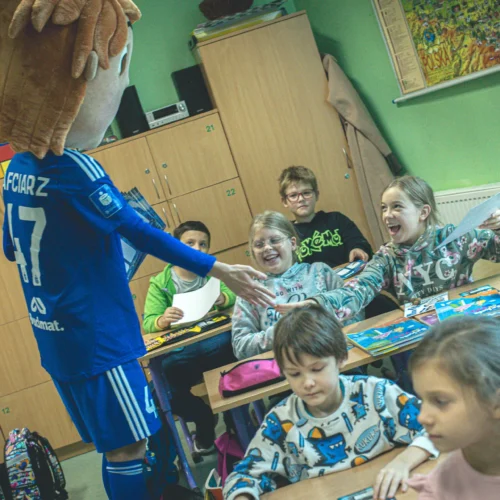 Na zdjęciu w klasie dzieci cieszy się z wizyty postaci w kostiumie sportowym, która wręcza im jakieś materiały.