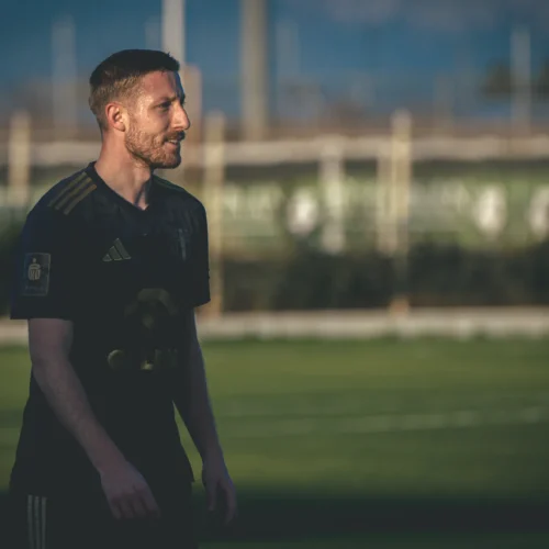Mężczyzna w ciemnej koszulce piłkarskiej stoi na boisku, z lekko uśmiechniętą twarzą, podczas treningu na tle zielonej murawy.