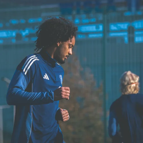 Mężczyzna w niebieskim stroju sportowym trenuje na boisku, biegnąc w kierunku lewej strony kadru.