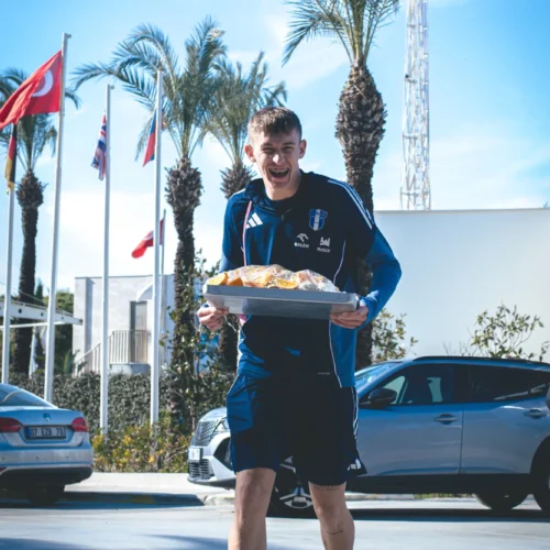 Młody mężczyzna w niebieskim stroju sportowym radośnie niesie tacę z jedzeniem na tle palm i flag.