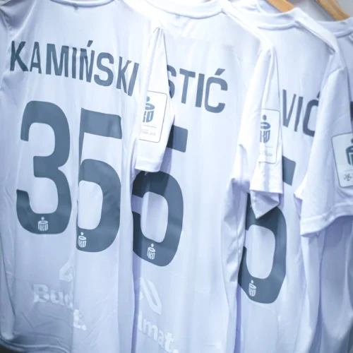 Widok kilku białych koszulek piłkarskich zawieszonych na wieszakach, z widocznymi nazwiskami zawodników oraz numerami na plecach.