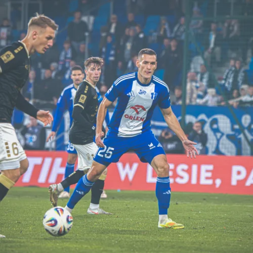 Na zdjęciu widać zawodników biorących udział w meczu piłki nożnej, z akcją rozgrywaną na boisku w tle.