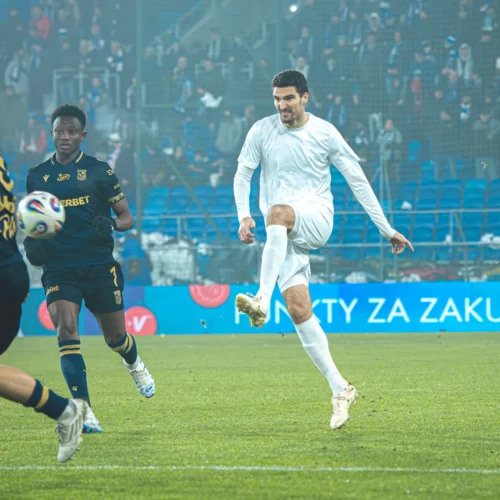 Na zdjęciu widać dwóch piłkarzy w strojach sportowych, którzy rywalizują na boisku, podczas gdy trzeci wykonuje techniczny ruch z piłką.