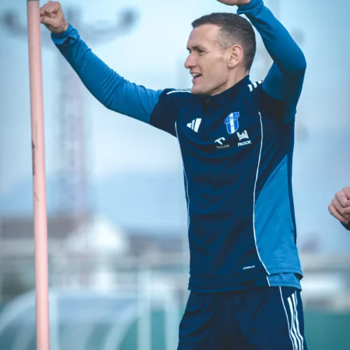 Na zdjęciu widać mężczyznę w niebieskiej odzieży sportowej, który radośnie unosi ręce w geście triumfu na boisku treningowym.