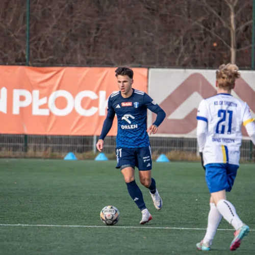 Na zdjęciu widać dwóch młodych piłkarzy podczas meczu na boisku, jeden w niebieskim stroju zmierza z piłką, a drugi w białym czeka na odpowiednią okazję.