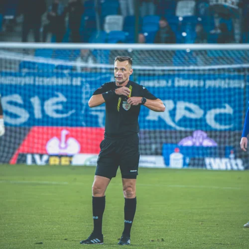 Na zdjęciu widać sędziego piłkarskiego wskazującego na coś na boisku, podczas meczu, w otoczeniu zawodników.