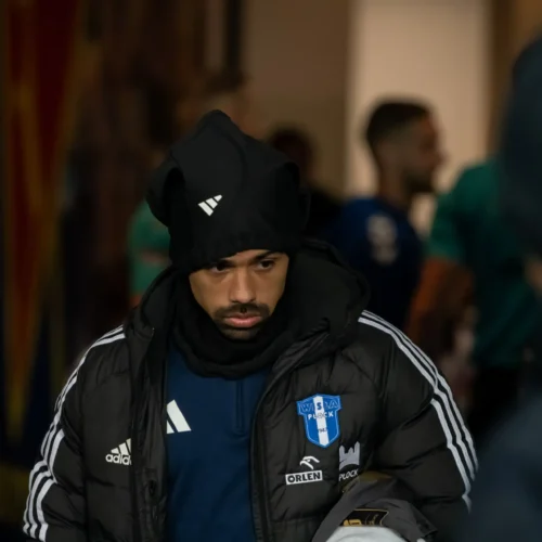 Na zdjęciu mężczyzna w kurtce z logo drużyny sportowej i czapce przechodzi przez korytarz.