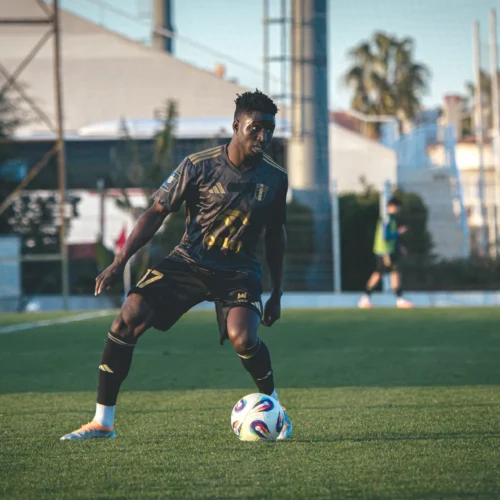 Na zdjęciu widoczny jest piłkarz grający w piłkę nożną na boisku, w strojach w odcieniach czerni i złota.