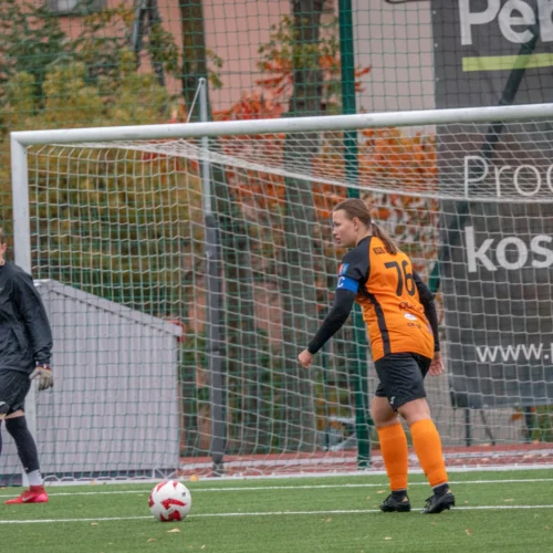 Na zdjęciu widoczna jest bramkarka w czarnym stroju oraz zawodniczka w pomarańczowej koszulce z numerem 76, obie na boisku piłkarskim, w trakcie meczu.