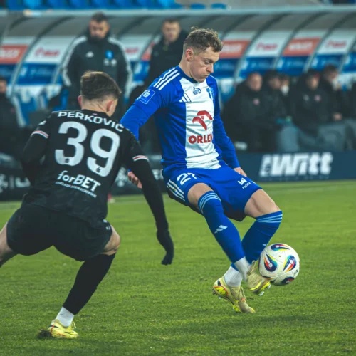 Na zdjęciu dwóch piłkarzy walczy o piłkę podczas meczu na zielonej murawie, jeden z nich ma na sobie niebiesko-białą koszulkę, a drugi czarną z numerem 39.