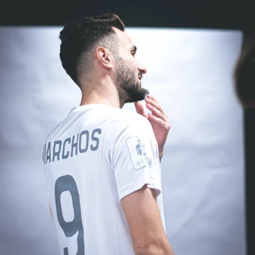 Mężczyzna w białej koszulce z numerem 9 i nazwiskiem "NARCHOS" stoi z lekkim uśmiechem, odwrócony w bok od obiektywu.