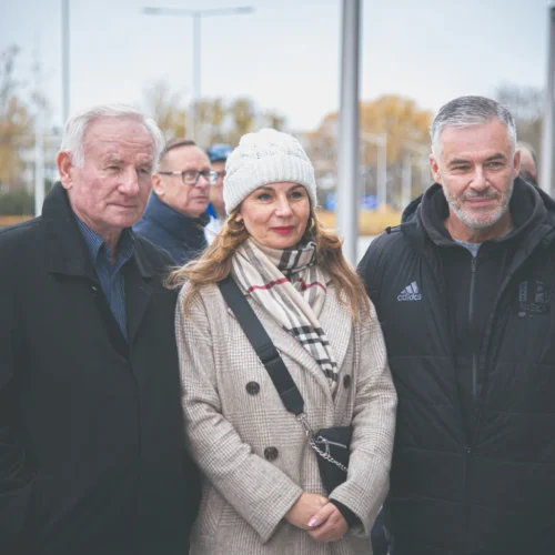 Na zdjęciu widoczna jest grupa trzech osób pozujących na zewnątrz w chłodny dzień, z widocznymi drzewami i budynkami w tle.
