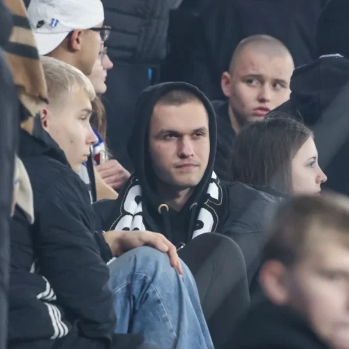 Na zdjęciu widać grupę kibiców na stadionie, skupionych na wydarzeniu sportowym, z wyraźnym napięciem emocjonalnym na ich twarzach.