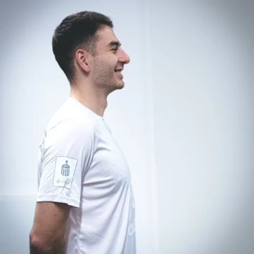 Mężczyzna w białej koszulce sportowej stoi w profilu, uśmiechając się.