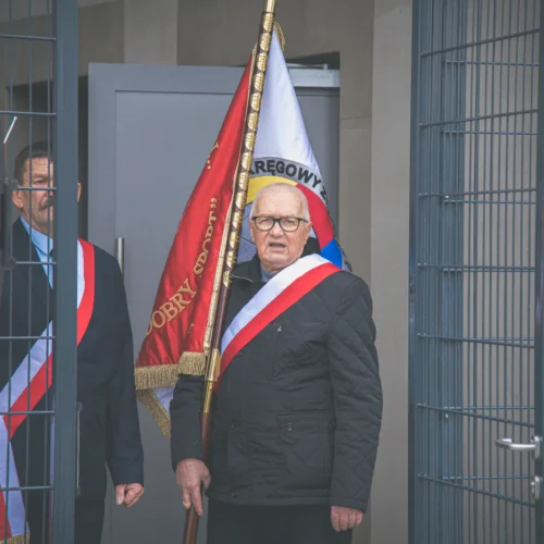 Na zdjęciu widoczni są dwaj mężczyźni w eleganckich strojach, z których jeden trzyma sztandar, podczas uroczystości.