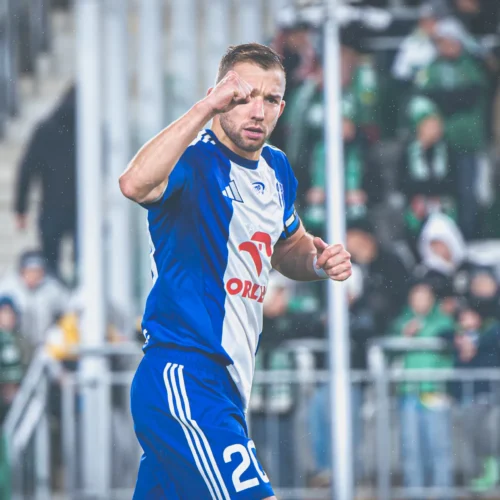 Na zdjęciu sportowiec w niebieskiej koszulce unosi pięść w geście triumfu podczas meczu piłkarskiego, a w tle widać zamożnych kibiców.