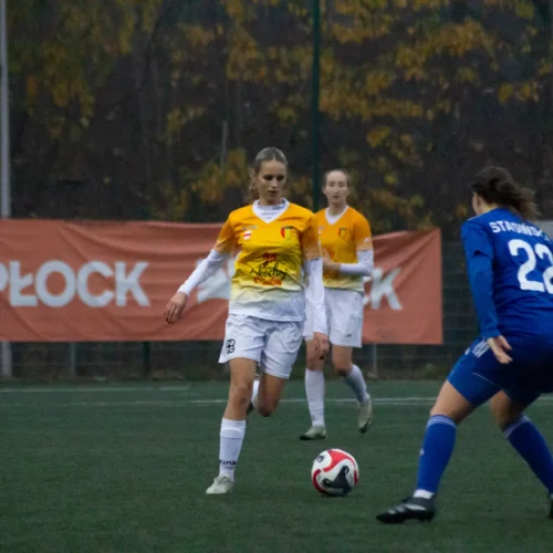 Na zdjęciu widoczna jest akcja meczu piłki nożnej kobiecej, gdzie zawodniczki w żółtych i niebieskich strojach rywalizują na boisku w pochmurny dzień.