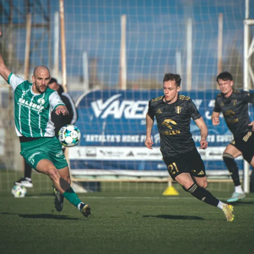 Na zdjęciu dwóch piłkarzy walczy o piłkę na boisku, a w tle widać bramkę oraz sędziów.